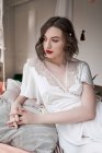 Preciosa mujer con labios rojos en vestido blanco mirando hacia otro lado mientras está sentada en el suelo junto al sofá - foto de stock