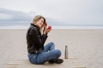 Vista lateral de la mujer rubia de moda en gorra negra y chaqueta de cuero comiendo manzana roja madura en la playa de arena - foto de stock