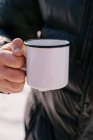 Abgeschnittenes Bild eines Mannes in warmer schwarzer Jacke mit weißem Emaille-Becher und heißem Tee, während er an einem Wintertag in Sibirien im Freien steht — Stockfoto