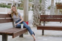 Вид сбоку жизнерадостной случайной женщины, сидящей на городской скамейке у моря в летний день и отводящей взгляд — стоковое фото