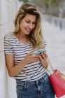Happy cheerful woman in casual striped shirt and jeans standing next to building on city street and using smartphone — Stock Photo