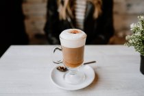 Обрізане зображення модної жінки, що сидить за столом зі склянкою смачної пінистої кави на столі — стокове фото