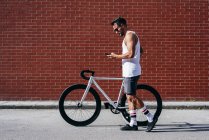 Bello ciclista di sesso maschile in abbigliamento sportivo e occhiali da sole utilizzando smartphone mentre in piedi con la bicicletta accanto al muro di mattoni rossi — Foto stock