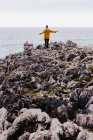 Vista posteriore della donna in felpa gialla con cappuccio che calpesta la riva pietrosa circondata da onde schiumose nella giornata nuvolosa grigia — Foto stock