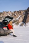Вид збоку жінки, що сидить на багажнику відкритого автомобіля і використовує телефон у засніженій долині з красивими скелями під час зимової поїздки — стокове фото