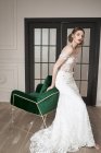 Barefooted slender bride in elegant white dress looking at camera while sitting on backrest of green armchair against black doors in spacious apartment — Stock Photo