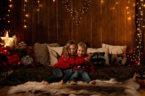 Entzückende kleine Mädchen und Jungen, die sich umarmen, als säßen sie in einem Raum voller Weihnachtsdekoration und schauen in die Kamera — Stockfoto