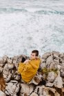 Dall'alto della donna in felpa gialla calda con cappuccio seduta da sola sulla riva rocciosa con onde schiumose nella giornata nuvolosa e guardando la fotocamera — Foto stock