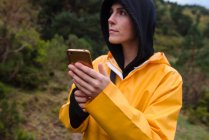 Задумчивая молодая женщина в тёмной толстовке и жёлтом плаще со смартфоном, смотрящая в лес — стоковое фото