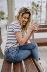 Вид сбоку на веселую случайную молодую женщину, пьющую из чашки кофе на вынос, сидящую на городской скамейке у моря в летний день — стоковое фото