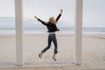 Visão traseira da mulher na moda em jaqueta preta saltando alegremente com os braços levantados no cais de madeira branca com oceano no fundo — Fotografia de Stock