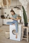 Chilling young barefoot woman having break and comfortably sitting on washing machine surfing mobile phone in kitchen — Stock Photo