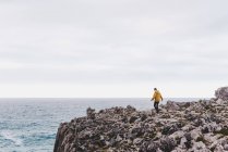 Seitenansicht einer Frau in gelbem Kapuzenpulli, die an einem grauen bewölkten Tag am steinigen Ufer von schäumenden Wellen umgeben ist — Stockfoto