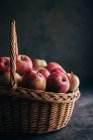 Frische rote Äpfel auf dunklem Tisch und in einem Weidenkorb auf dunklem Hintergrund — Stockfoto