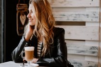 Боковой вид на длинноволосатую модную красивую блондинку, сидящую в кафе и пьющую из стакана вкусного пенного кофе — стоковое фото