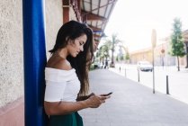 Щаслива молода жінка з довгим волоссям спирається на стіну дивиться на мобільний телефон — стокове фото