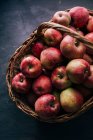 Frische rote Äpfel auf dunklem Tisch und in einem Weidenkorb auf dunklem Hintergrund — Stockfoto