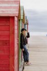 Trendy long haired blonde woman in black cap and leather jacket smiling brightly at camera and leaning on wall of wooden beach cabins — Stock Photo