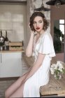 Hermosa mujer tierna en bata de seda blanca tocando el cabello y reflexionando mientras está sentado en la mesa junto a las flores en el apartamento - foto de stock