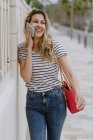 Счастливая веселая женщина в повседневной полосатой рубашке и джинсах стоит рядом со зданием на городской улице и разговаривает по смартфону — стоковое фото