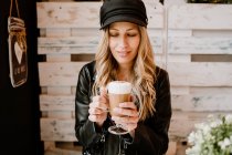 Длинноволосая модная женщина держит стакан вкусного пенного кофе в кафе — стоковое фото