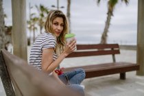Vista lateral de la alegre mujer casual con una taza de café para llevar sentado en el banco de la ciudad en primera línea de mar en el día de verano - foto de stock