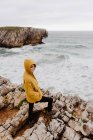 Vista posteriore della donna in felpa calda gialla in piedi da solo sulla riva rocciosa con onde schiumose nella giornata nuvolosa — Foto stock