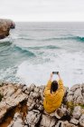 Back view of short haired woman in yellow sweatshirt sitting on rocky seashore and taking selfie on mobile phone — Stock Photo