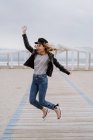 Trendfrau in schwarzer Jacke springt fröhlich mit erhobenen Armen auf weißem Holzsteg mit Ozean im Hintergrund — Stockfoto