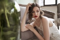 Preciosa mujer con labios rojos en vestido blanco mirando hacia otro lado mientras está sentada en el suelo junto al sofá - foto de stock
