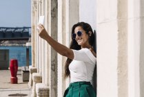 Attractive young female in trendy outfit posing for selfie while standing on bright background — Stock Photo