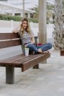 Вид збоку впевнена жінка сидить на міській лавці на набережній в літній день, дивлячись на камеру — стокове фото
