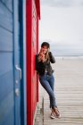 Donna alla moda in berretto nero e giacca di pelle che mangia una mela rossa mentre si appoggia su cabine da spiaggia in legno — Foto stock