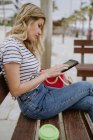 Side view of woman sitting on street bench at seafront with disposable cup of coffee and browsing internet on tablet — Stock Photo