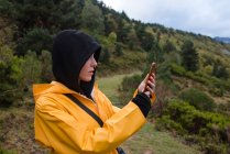 Mulher com capuz e capa de chuva amarela navegando smartphone ao ar livre — Fotografia de Stock