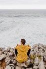 Visão traseira do viajante com capuz quente amarelo sentado sozinho na costa rochosa olhando para ondas espumosas no dia nublado — Fotografia de Stock
