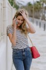 Счастливая веселая женщина в повседневной полосатой рубашке и джинсах стоит рядом со зданием на городской улице и разговаривает по смартфону — стоковое фото