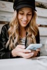 Стильная длинноволосая блондинка в черной кожаной куртке и модной кепке, улыбающаяся во время сёрфинга мобильного телефона в руках за столом в кафе — стоковое фото