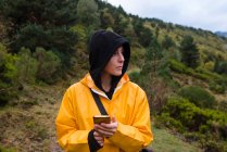 Attraktive Frau in Kapuzenpulli und gelbem Regenmantel surft im Smartphone — Stockfoto