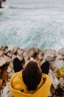Back view of traveler in yellow warm hoodie sitting alone on rocky shore looking at foamy waves on cloudy day — Stock Photo