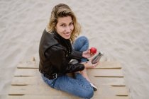 Сверху модная блондинка в кожаной куртке ест красное спелое яблоко на песчаном пляже, смотрит в камеру — стоковое фото