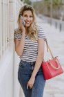 Happy cheerful woman in casual striped shirt and jeans standing next to building on city street and talking on smartphone — Stock Photo