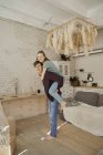 Side view of smiling handsome man giving piggyback ride to cheerful female in light cozy living room — Stock Photo