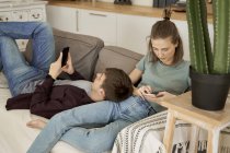 Спокойный вдумчивый молодой человек и женщина, лежащие на уютном мягком диване и серфинг мобильных телефонов дома — стоковое фото