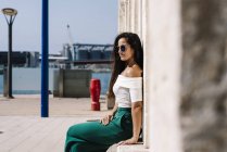 Side view of a young beautiful hispanic woman with sunglasses sitting on a stone bench against wall looking away — Stock Photo
