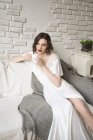 Потрясающая взрослая женщина смотрит в сторону и мечтает, сидя на диване и наслаждаясь горячим напитком в светлой современной гостиной — стоковое фото