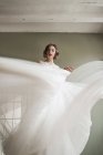 Снизу великолепная расслабленная женщина танцует и машет светлой прозрачной тканью белого свадебного платья во время подготовки к событию в квартире — стоковое фото