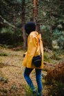 Seitenansicht einer Frau mit schwarzer Kapuze und gelbem Regenmantel, die mit der Kamera in der Tasche im Kiefernwald unterwegs ist — Stockfoto