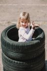 Сверху счастливая очаровательная маленькая девочка, стоящая в стопке автомобильных шин, веселясь и играя на открытом воздухе в летний день — стоковое фото