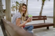 Вид сбоку женщины с чашкой кофе, сидящей на городской скамейке у моря в летний день — стоковое фото